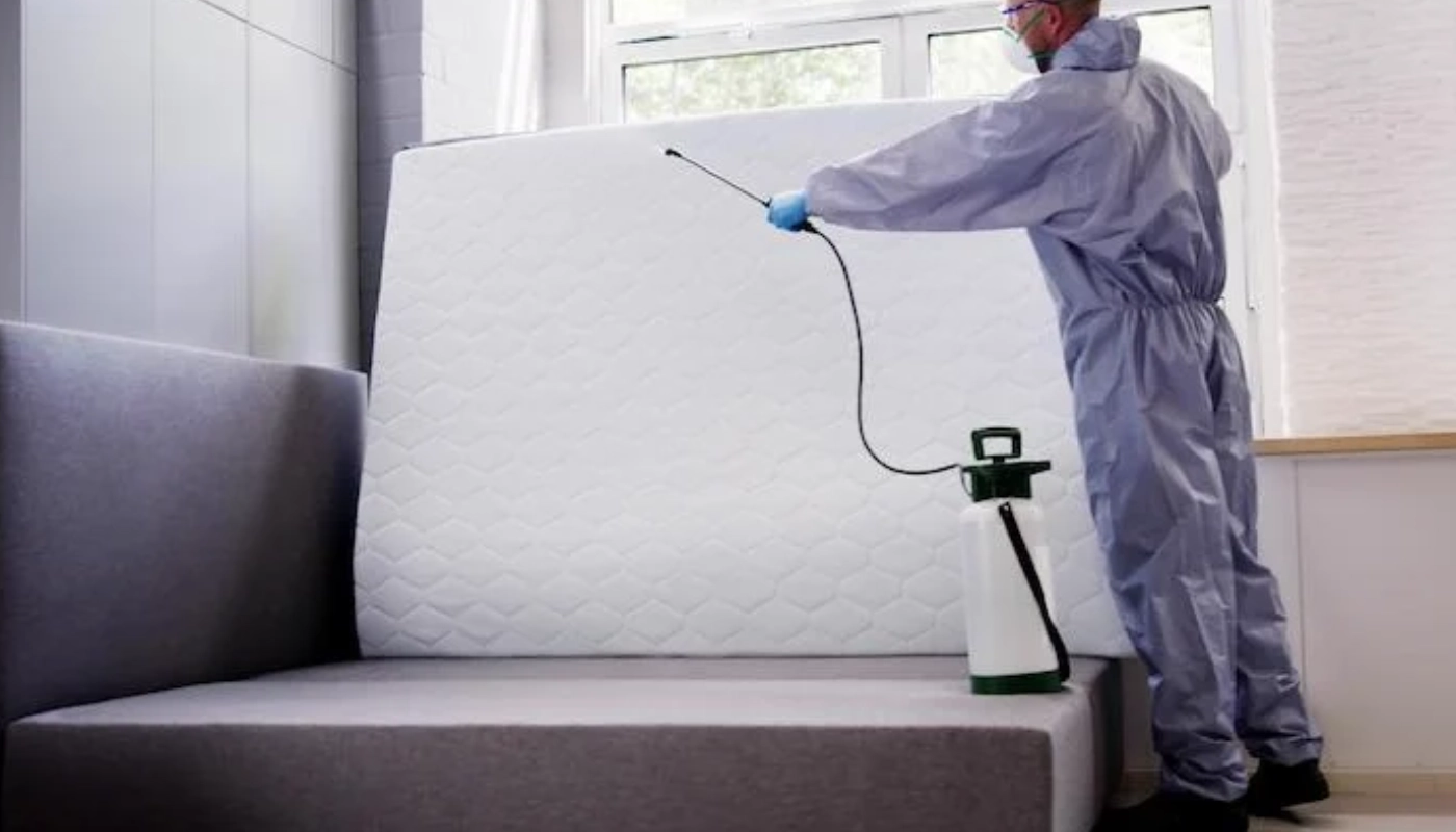 Technicien en combinaison de protection appliquant un traitement professionnel contre les punaises de lit sur un matelas relevé, à l’aide d’un pulvérisateur.
